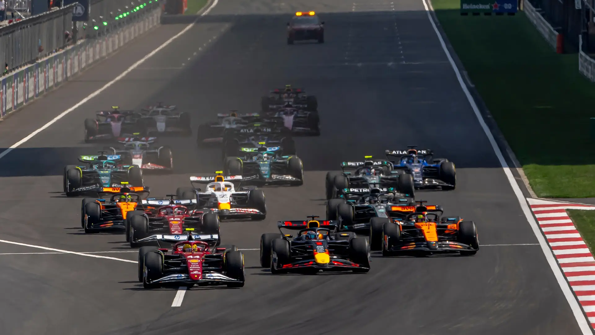 A group of Formula 1 cars racing side by side on a wide track during the start of a race, with colorful liveries and a safety car visible in the distance.