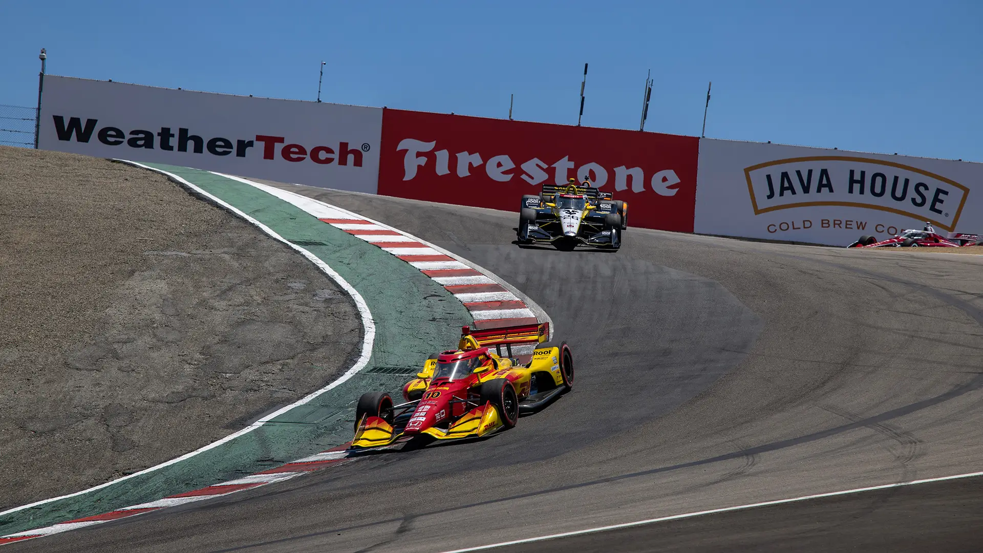 A red and yellow IndyCar leading through a sharp uphill turn on a racetrack, followed by a black and yellow car, with sponsor banners for WeatherTech, Firestone, and Java House visible on the barriers.