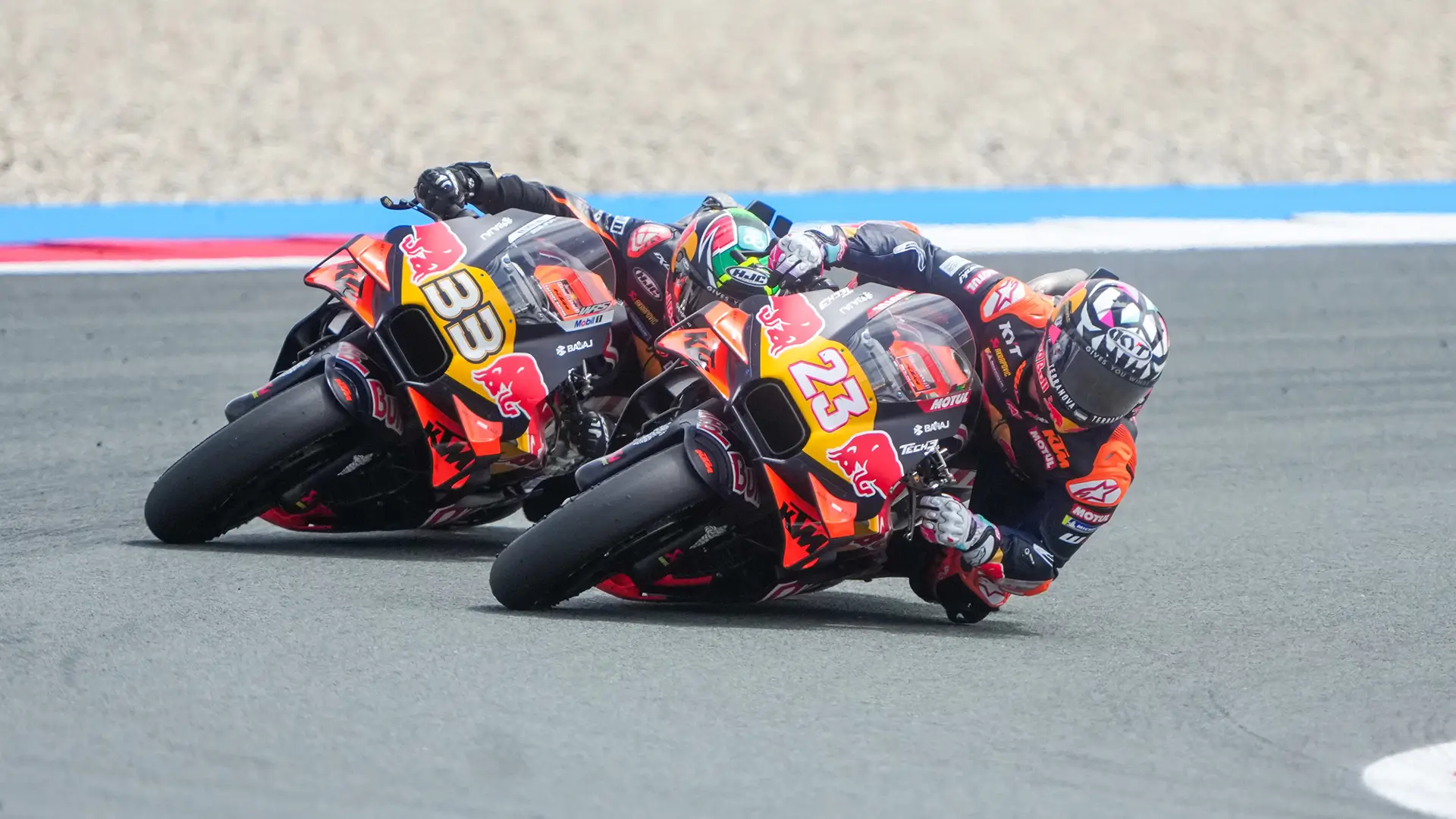 Two MotoGP riders leaning sharply into a corner on a racetrack, riding orange and black motorcycles with visible numbers 33 and 23.