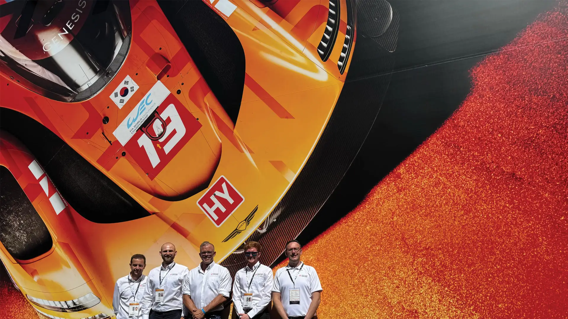Five people in branded white shirts stand in front of a large display featuring an orange Le Mans prototype race car with number 19 and WEC markings.