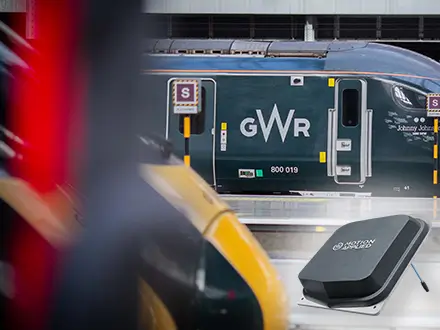 Great Western Railway (GWR) train carriage at a station platform, with the GWR logo visible on the side. Out-of-focus trains partially frame the foreground, and an electronic ticket validator is positioned in the lower right corner.