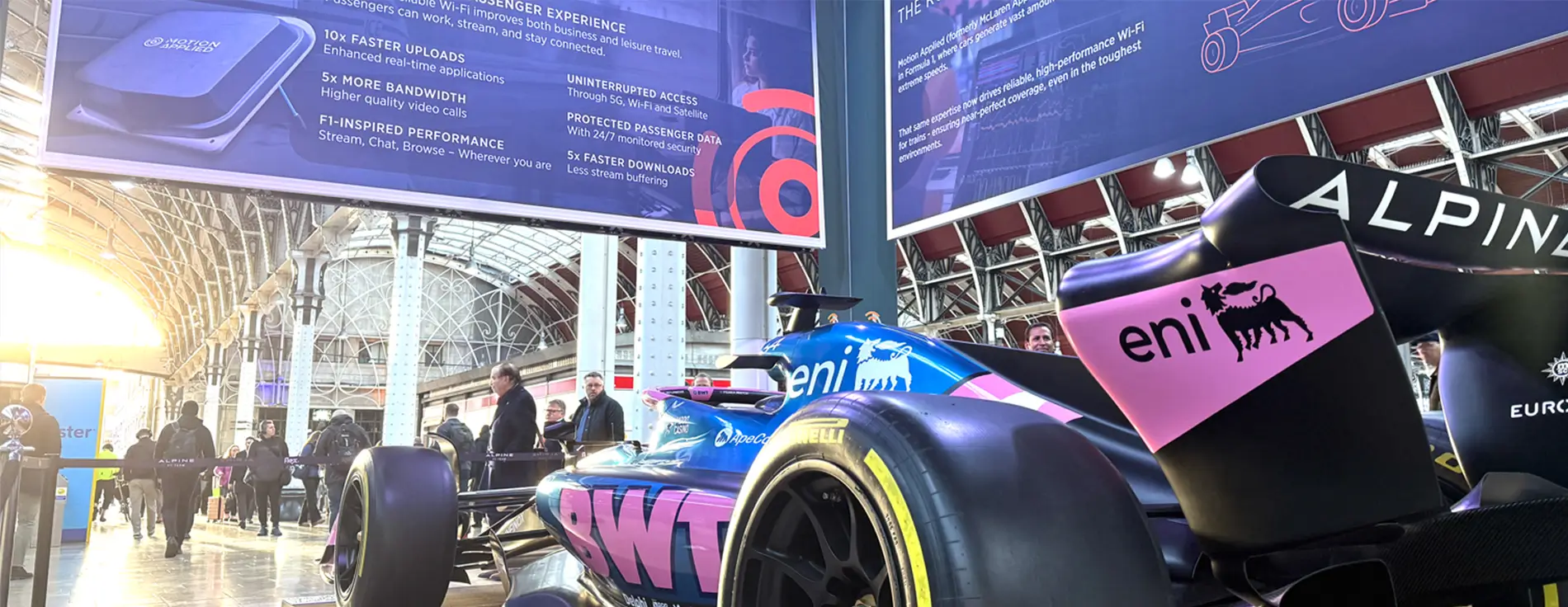 A blue and pink Alpine Formula 1 race car displayed indoors at a train station concourse, with large promotional banners overhead advertising high-speed Wi-Fi features. Sunlight streams through the glass roof, and people walk around the exhibit in the background.