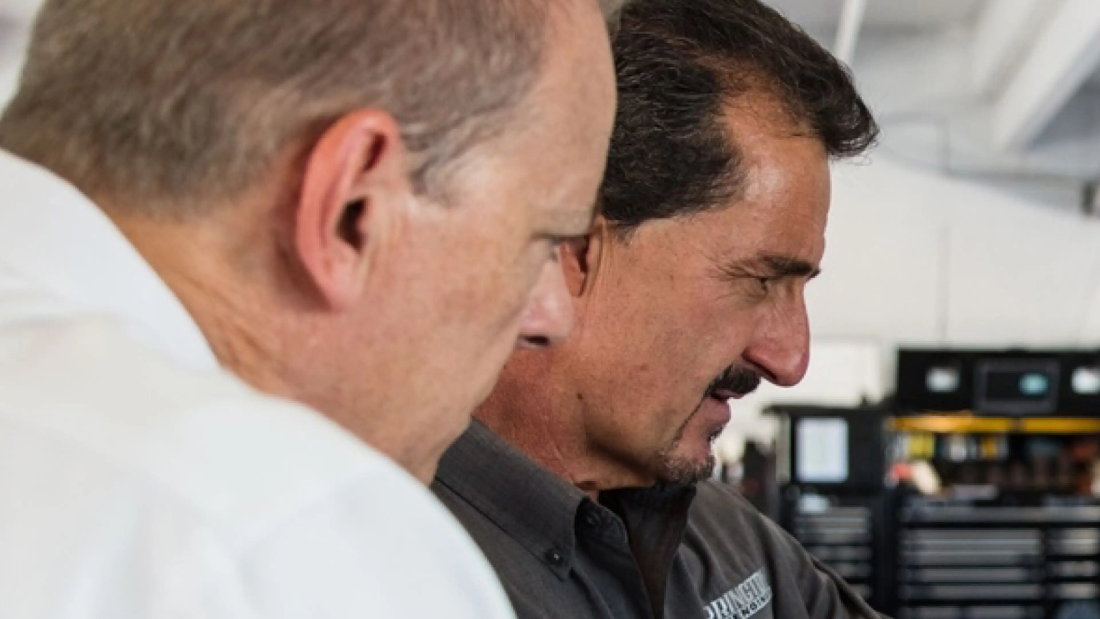 Close-up of two people working together in a workshop or technical support environment, focusing on equipment or a task off-camera, with tools and storage units visible in the background.